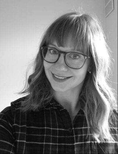 Black and white picture of a young woman with light, shoulder-length hair and a fringe. She is wearing a large pair of glasses and a checked shirt. She is smiling into the camera.